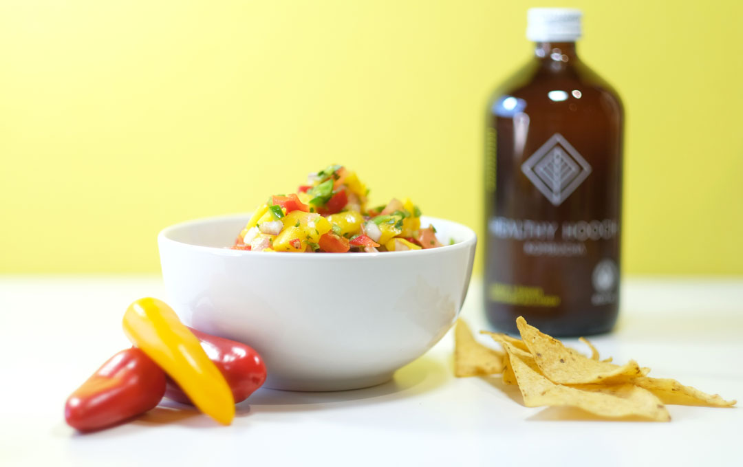 mango salsa next to peppers, tortilla chips and a bottle of kombucha